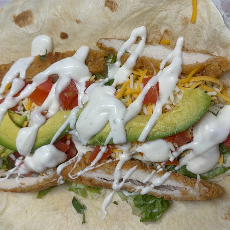 A chicken strip burrito with avocado, tomatoes, cheese, lettuce, and white sauce on a tortilla.