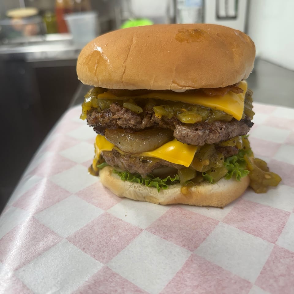 A double cheeseburger with green chili, onions, and lettuce on a red and white checkered paper.