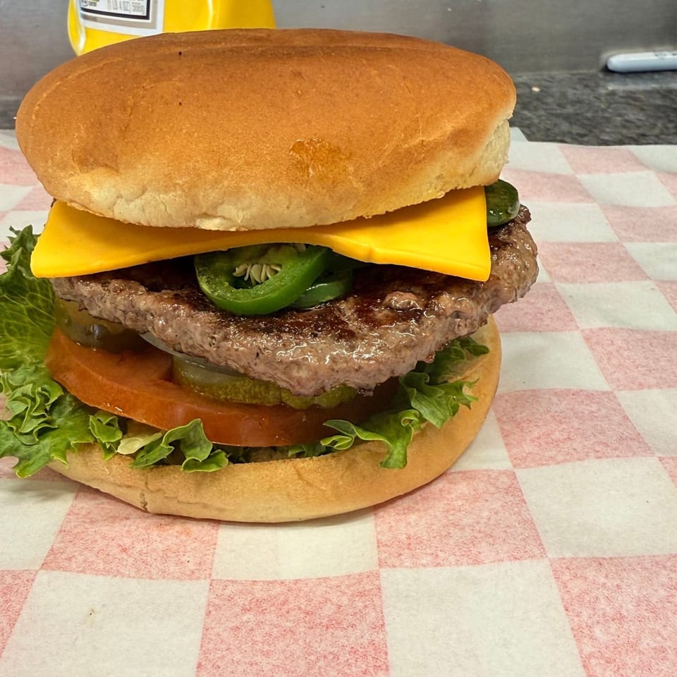 A cheeseburger with lettuce, tomato, pickles, and jalapeños on checkered paper, with mustard in the background.