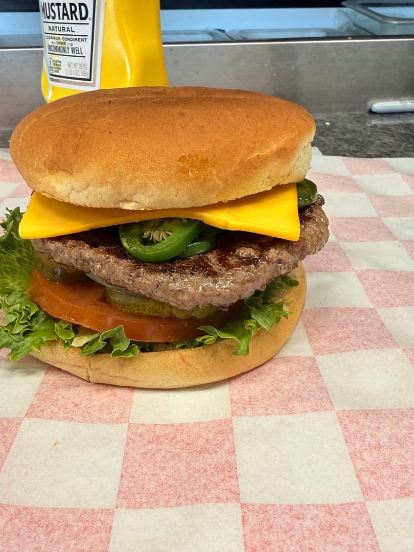 A cheeseburger with lettuce, tomato, pickles, and jalapeños on checkered paper, with mustard in the background.