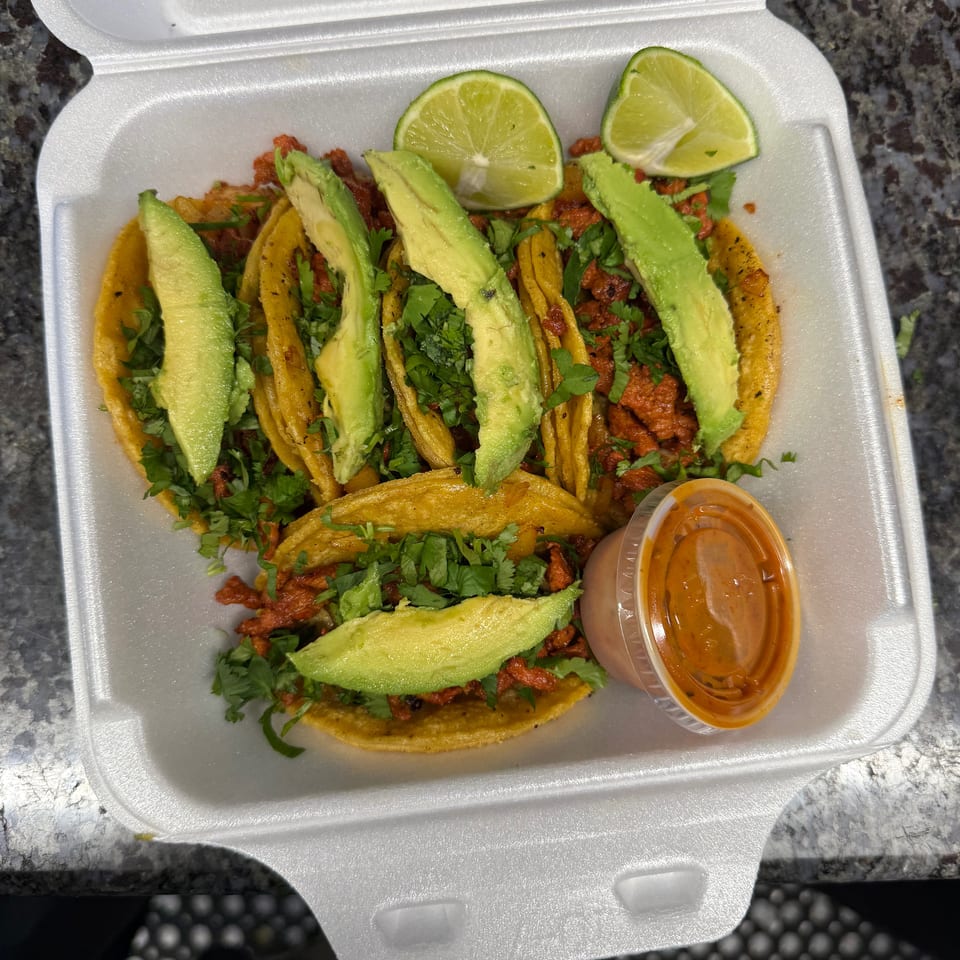 Five tacos with meat, cilantro, and avocado slices, with lime wedges and a side of sauce.